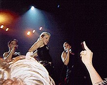 Right profile of a blond woman, flanked by two other woman who are singing in a microphone. The blond woman wears a black t-shirt and her hair is tied in a bun. Behind them, lights flash from above. The hand and head of a person is visible below.