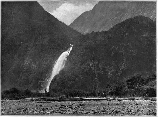 A waterfall coming down the middle of a bush-clad hill, with another hill in the background and a rocky flat plain in the foreground