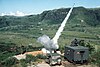 Two men watch as a small rocket roars off its launch rail next to a Jeep and small truck.