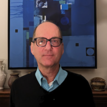 Photograph of Greg Hewett, a white, balding man with thick-rimmed glasses. He is wearing a black sweater over a light blue button-down shirt. He appears in front of a blue geometric painting and various pieces of pottery.