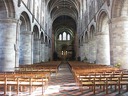 Stone columns supporting arches form the side walls of the long hall, and wooden chairs are set up on either side of a central aisle.