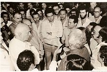 Fotografia em preto e branco, mostra Tancredo Neves, o candidato civil às Diretas, em 1984, recendo documento entregue por trabalhadores, com a presença de muitas personalidades.