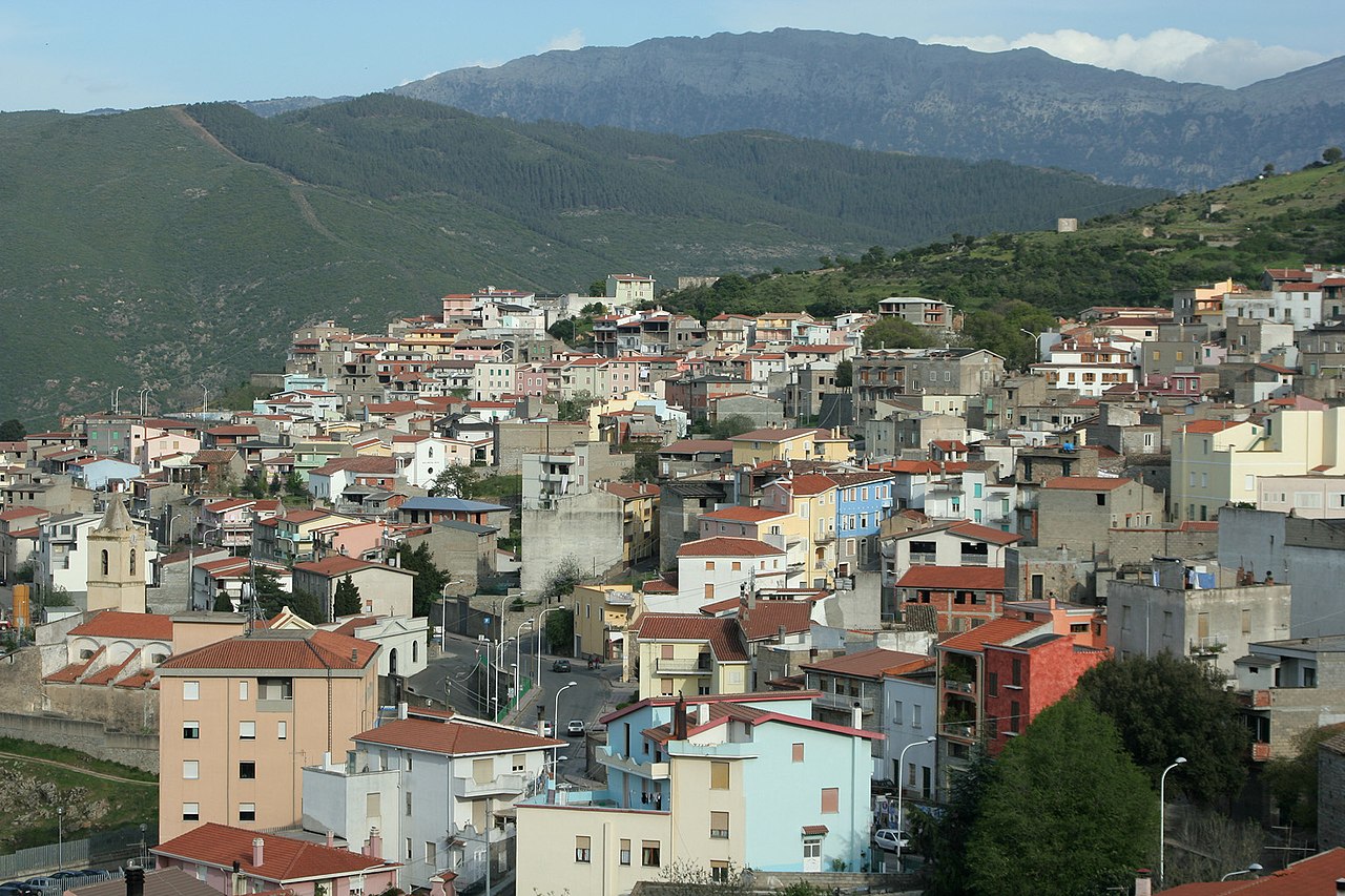 The village of Orgosolo with green hills in the background and the buildings are almost all multi stories with reddish roofs and they are clustered close together