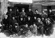 Photo noir et blanc d'une vingtaine d'hommes en chapka posant devant un bâtiment, certains tiennent un fusil