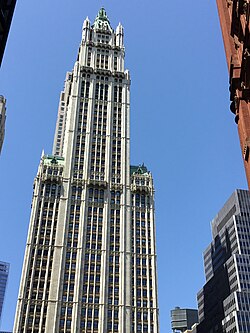 Color photo of the Woolworth Building, a skyscraper, with trees in the foreground and a shorter building to the left