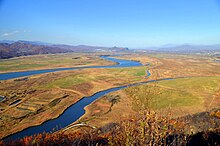 Photographie depuis une hauteur d'une vallée large et plate, avec deux bras d'une même rivière au centre, avec au fond des montagnes.