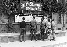 Черно-бяла историческа снимка на няколко мъже, някои с нацистки ленти, четящи билборд във вестник.