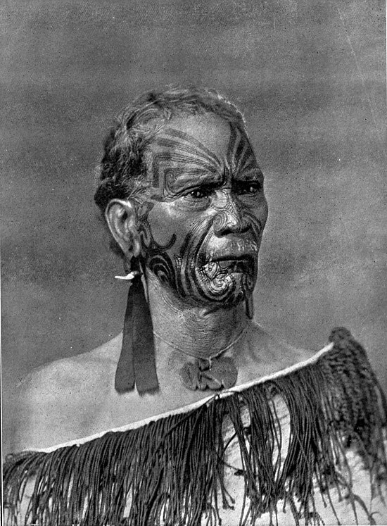 An elderly Māori man with full facial moko, two feathers in right ear, hei tiki, and fringed cloak.