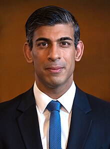 Sunak facing frontward, with combed over black hair and a dark blue suit in front of a brown background.