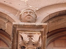 Photo couleur présentant le chapiteau d'une colonne. Décoré de feuillages, il est surmonté d'une tête à la jointure des arcs brisés qui en partent.