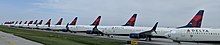 Many in line airplanes with the Delta Air Lines logo on the tail, parked on pavement behind a fence.