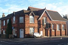 A dark red brick hall on a corner site, with adjoining houses in an identical style. The hall, partly obscured by a traffic light, has a pointed-arched entrance with a wooden door below a rectangular window frame with three lancets. To the right of the door, there are three plain windows between brick buttresses. Above the entrance is a steep roof perpendicular to the main roof.