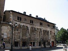 Photographie représentant un bâtiment doté de voûtes romanes, situé en ville devant une place.