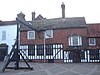 A tile-hung and timber-framed building with an uneven tiled roof and a prominent bay window to the right. There is a tall chimney-stack on the roof, and a gallows extends from the ground in front to just below the eaves.