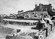 In a walled enclosure, there are several tents and an empty rectangle surrounded by tens of mounted knights. A castle rises in the background.