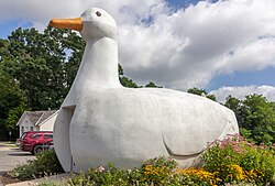 A building which looks like a large white duck with an orange beak. The duck appears to be sitting on the ground. There is a doorway in the front, below the head.