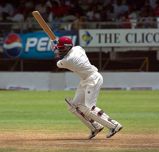 A man wearing white cricket clothes and maroon helmet plays a shot. He is standing on a cricket pitch, and the seating area of the ground is vi