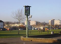 A photograph of an urban park with houses and flats in the distance. The park large, and mostly grass. A football goal is in the centre of the park, and a pub sign is in the foreground.