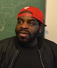 Hanif Willis-Abdurraqib pictured in front of a map of Ohio. He wears a black shirt and jacket and a red baseball cap, turned backwards, and looks off-camera.