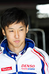 A man in his late 20s wearing a white and blue jacket with sponsors logos on both sides is looking to the extreme left of the camera