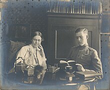 Margaret Maltby sits drinking tea at a table with Philip Randolph Meyer. The interior is decorated with fine objects. in the background is a wardrobe. Maltby has her hair back in a bun and leans on to one side, resting her head on her hand while gazing at the camera. Meyer is young and dressed in a World War I army uniform. He gazes downward at an open book as if reading.