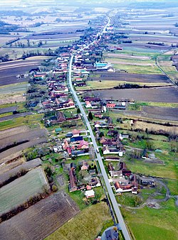 Aerial photograph of the village. There are houses on either side of a central road and fields beyond the houses