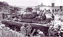 Photo noir et blanc. Au premier plan un char sur une plage en appui des troupes débarquant. Au loin une île.