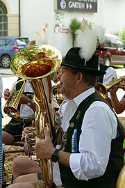 Foto des Oberkörpers eines Mannes, der in ein Blechblasinstrumen bläst. Er trägt einen grünen Hut mit Federn. Im Hintergrund sind weitere Musiker zu sehen