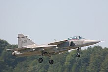 Grey jet fighter on take off facing right of photo against a background of green trees and blue sky. Its landing gear is still down.