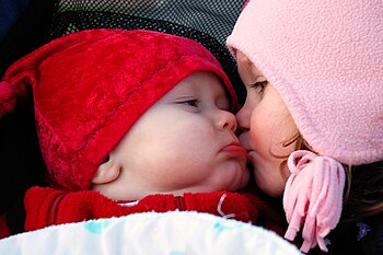 English: A young girl kisses a baby on the cheek.