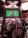 A silver motorcycle with black tires sits on a podium. A TV screen with a green preview displayed sits behind the motorcycle with a large X above it and a picture of Wolverine below.