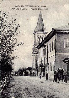 Czarno-białe zdjęcie z XIX/XX w. przedstawiające ulicę o nierównej nawierzchni, przy której chłopcy w mundurkach przed piętrowym gmachem, na dalszym planie wieża kościelna
