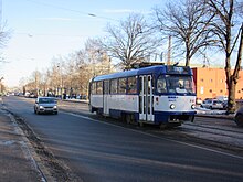 Seitliche Farbfotografie einer blau-weißen Straßenbahn, die über eine Kurve in eine Allee mit alten Gebäuden fährt. Rechts hinten ist ein weißes Gebäude mit rundem Mittelteil und grünem Dach sowie Ornamenten.