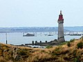 Phare blanc et rouge au sommet ; au premier plan, herbe jaunie ; en arrière plan, des rochers et la mer