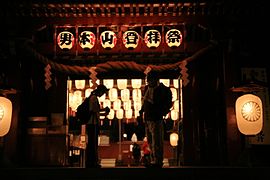 Photo couleur de deux personnes devant la porte ouverte d'un bâtiment d'un sanctuaire éclairé par des lampions, à la nuit tombée.