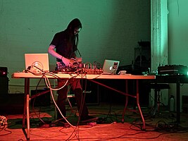 Akita stands behind a folding table covered in laptops and cords with green lighting on the walls behind.