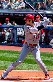 Un joueur de baseball, batte en mains, en tenue grise et casque rouge, s'apprête à frapper.