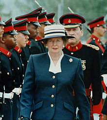 Thatcher walking in front of a line of military personnel, who are a mixture of races. Thatcher has a stern expression, dressed in a navy-coloured suit with buttons and a white hat.