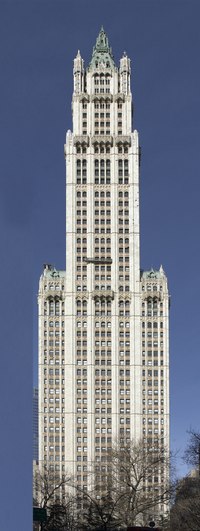 Color photo of the Woolworth Building, a skyscraper, with trees in the foreground and a shorter building to the left