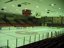 The image shows an empty Walter Brown Arena as viewed from the entrance to the arena behind the home net. The ice surface is empty. The image has a blue hue.