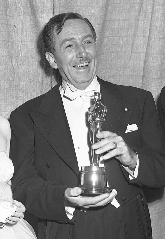 A black and white photograph of Walt Disney standing, holding an Academy Award.
