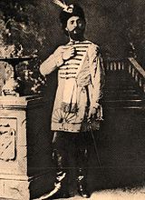 Монохромная фотография стоящего мужчины в кожаных книгах, тунике до колен и шляпе с перьями