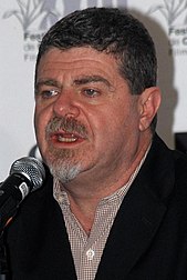 A man with dark gray hair talking into a microphone, looking beneath the camera