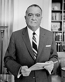 Photographie en noir et blanc de J. Edgard Hoover en costume portant un document.