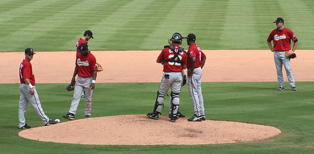 Seven men wearing red baseball jerseys, gray pants, and black caps are standing on a baseball diamond's infield; three are talking together on the mound.