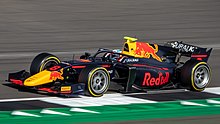 Une monoplace de Formule 2 aux couleurs Red Bull roulant sur un circuit avec des vibreurs verts.