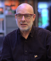 An old man wearing glasses and a dark blue shirt is interviewed