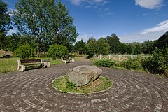 Kamień, wokół niego wyłożona kostka i ławki oraz drzewa w parku
