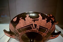 Pederastic couples. Boy at centre is holding an oenochoe in his left hand and giving a kylix to a person on a couch in his right hand. Attic kylix. Around 460-450 BCE Coupe - MSR - Rituels Grecs - AGER inv G 467.jpg