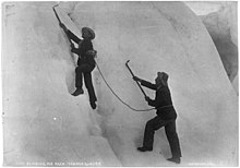 Mannering steht in einer Eiswand, ein Seil verbindet ihn mit dem tiefer stehenden Dixon. Beide tragen einfache Bergsteigerkleidung und Eispickel, aber keine Steigeisen.